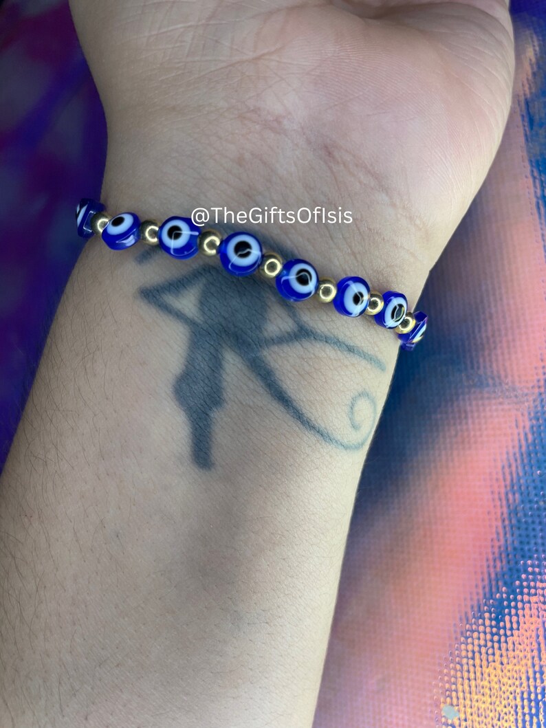 May include: A blue and gold evil eye bracelet with gold beads. The bracelet is on a person's wrist. @TheGiftsOfIsis