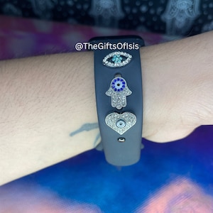 May include: A black wristband with three charms: a blue evil eye, a silver hamsa hand with blue accents, and a silver heart with a blue evil eye. The charms are adorned with rhinestones. The wristband is on a person's wrist. The text "@TheGiftsOfIsis" is visible in the image.
