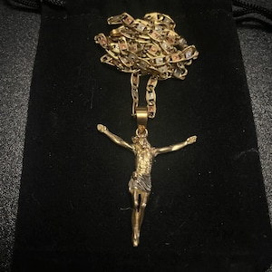 May include: A gold-coloured necklace featuring a pendant of a figure with outstretched arms. The chain is a mix of gold, silver, and rose gold links. The necklace is displayed on a black velvet pouch.