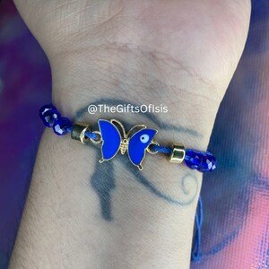 May include: Blue and gold butterfly charm bracelet with a blue evil eye charm. The bracelet is made with blue glass beads and a blue cord. The bracelet is on a person's wrist. @TheGiftsOfIsis