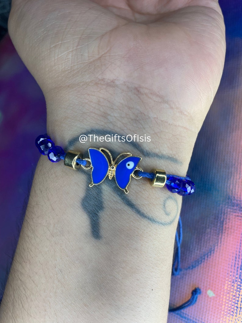 May include: Blue and gold butterfly charm bracelet with a blue evil eye charm. The bracelet is made with blue glass beads and a blue cord. The bracelet is on a person's wrist. @TheGiftsOfIsis