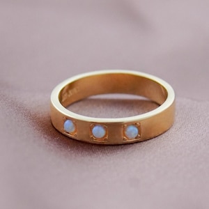 Puede incluir: Un anillo de oro con tres pequeñas piedras de ópalo cuadradas. El anillo tiene una banda plana y un acabado mate. Los ópalos son de un color blanco lechoso pálido. El anillo se muestra sobre una tela suave de color malva.