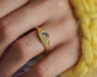 May include: A gold ring with a purple gemstone set in a square bezel. The ring is worn on a finger with a light skin tone.