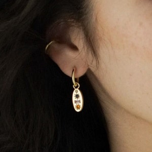 May include: A gold hoop earring featuring a pendant with an oval shape. The pendant is decorated with small gemstones in black, green, and orange. The earring is worn on an ear, with dark hair in the background.
