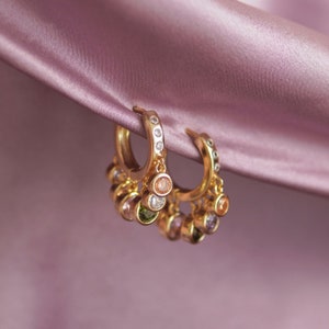 May include: A pair of gold hoop earrings with dangling charms featuring colourful gemstones. The earrings are displayed on a pink satin background.