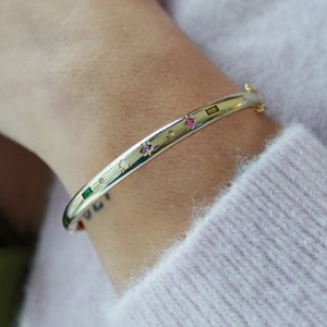 Op de afbeelding: Een gouden armband met een rij kleurrijke edelstenen, waaronder een roze, groene en witte steen.