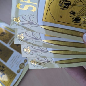 May include: A stack of four gold foil stickers with a floral design. The stickers have a white background and a gold border. The text "Spring" is visible on the top sticker.