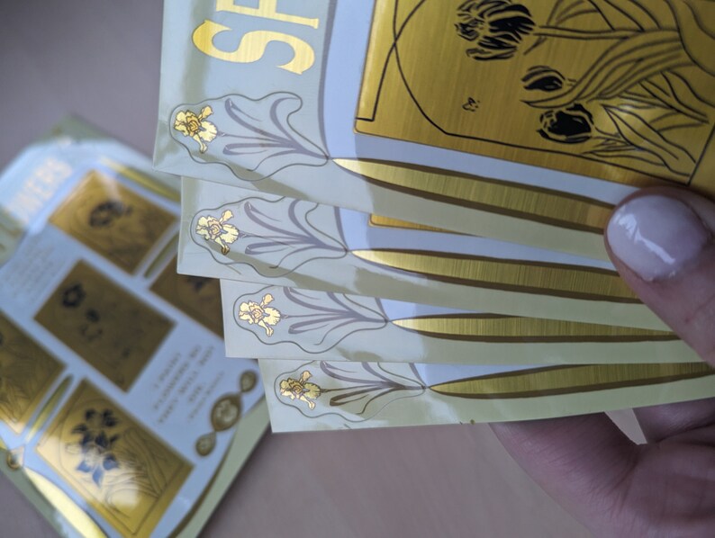 May include: A stack of four gold foil stickers with a floral design. The stickers have a white background and a gold border. The text "Spring" is visible on the top sticker.