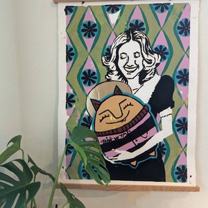 May include: A colorful print featuring a woman with long brown hair holding a cartoon character with a pink and black striped shirt. The background is a green and pink geometric pattern.