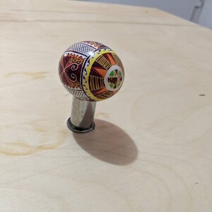 May include: A decorative Easter egg with intricate patterns in shades of red, brown, yellow, and green, mounted on a silver metal stand.