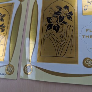 May include: Gold-toned sticker sheet with a floral design. The sheet features a black and white illustration of a lily flower with a gold background. The text "FLOWERS OF THE GARDEN" is also included on the sheet.