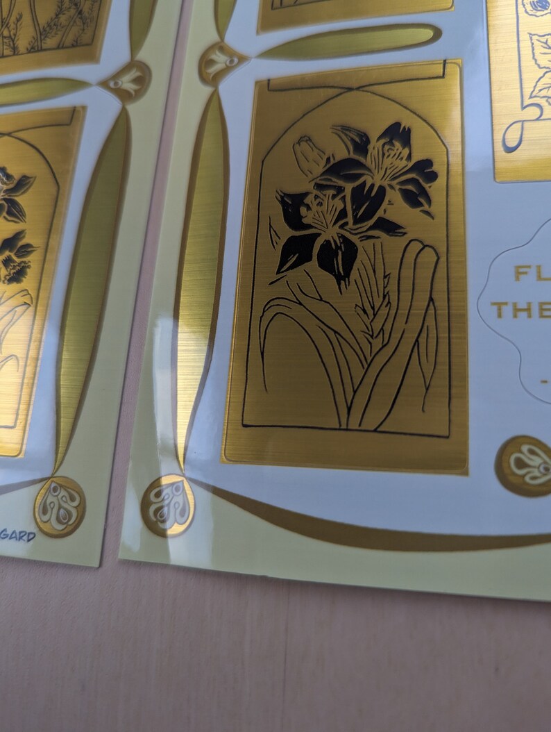 May include: Gold-toned sticker sheet with a floral design. The sheet features a black and white illustration of a lily flower with a gold background. The text "FLOWERS OF THE GARDEN" is also included on the sheet.