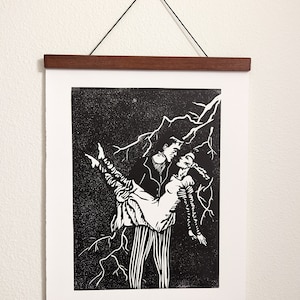 May include: Black and white linocut print of a couple dancing in a stormy night. The man is holding the woman in his arms, and they are both smiling. The print is framed in a dark wood frame.