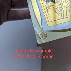 May include: A close-up of a gold-colored sticker with a floral design. The sticker has a small fold in the corner.  Grade B Example. Small fold in corner.