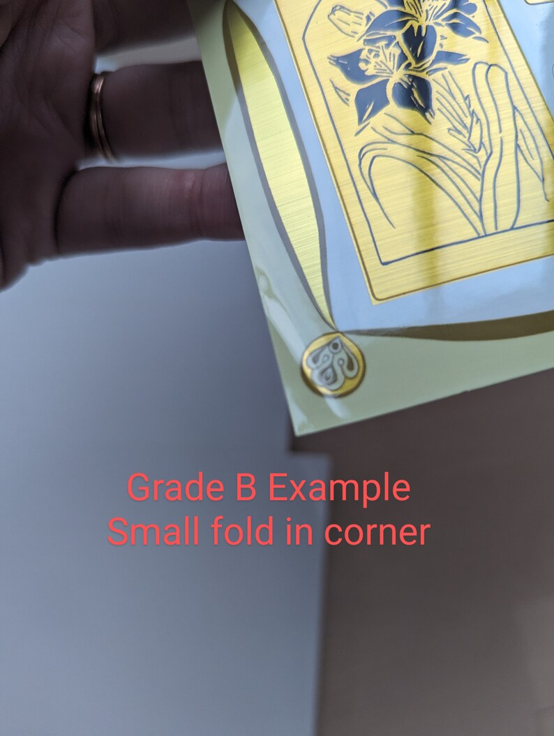 May include: A close-up of a gold-colored sticker with a floral design. The sticker has a small fold in the corner.  Grade B Example. Small fold in corner.