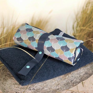 Puede incluir: Una manta de picnic enrollada con un patr&oacute;n geom&eacute;trico azul y blanco. La manta est&aacute; encima de un material azul oscuro, similar al fieltro, con una correa y dos botones turquesa.