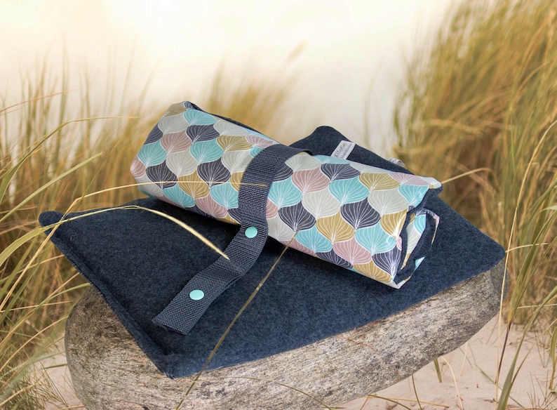 Puede incluir: Una manta de picnic enrollada con un patr&oacute;n geom&eacute;trico azul y blanco. La manta est&aacute; encima de un material azul oscuro, similar al fieltro, con una correa y dos botones turquesa.