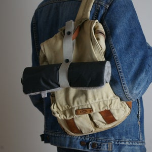 Puede incluir: Una persona lleva una chaqueta vaquera azul con una mochila de lona beige con una correa de cuero marr&oacute;n y una bolsa enrollable negra y gris adjunta. La mochila tiene un logotipo "Camel" en la parte delantera.