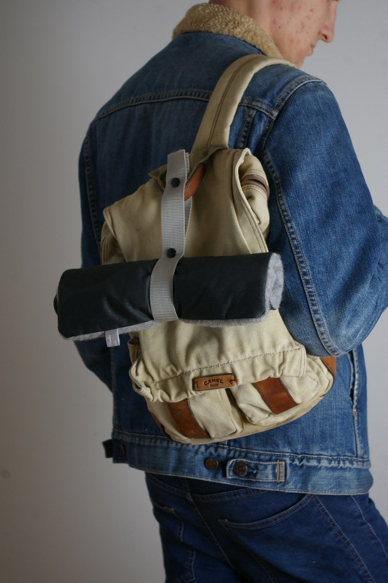 Puede incluir: Una persona lleva una chaqueta vaquera azul con una mochila de lona beige con una correa de cuero marr&oacute;n y una bolsa enrollable negra y gris adjunta. La mochila tiene un logotipo "Camel" en la parte delantera.