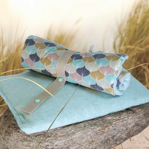 Puede incluir: Una toalla de playa enrollada con estampado de color azul verdoso y marr&oacute;n con una correa gris y botones turquesa. La toalla est&aacute; sobre una estera de playa de microfibra azul verdoso.