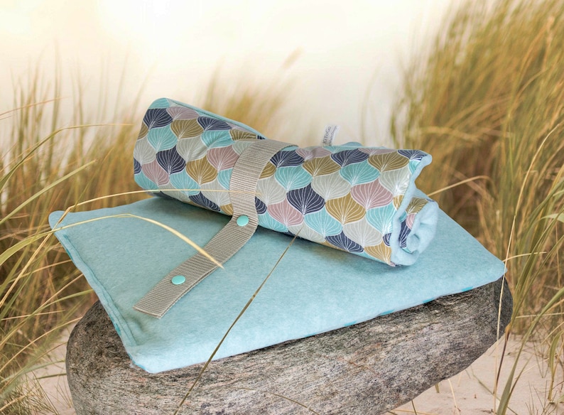 Puede incluir: Una toalla de playa enrollada con estampado de color azul verdoso y marr&oacute;n con una correa gris y botones turquesa. La toalla est&aacute; sobre una estera de playa de microfibra azul verdoso.