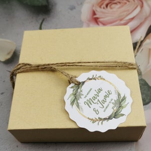 May include: A brown cardboard box with a white circular tag tied with twine. The tag has a gold circle with green leaves and the text "The Wedding Maria & Jamie Saturday 30.12."