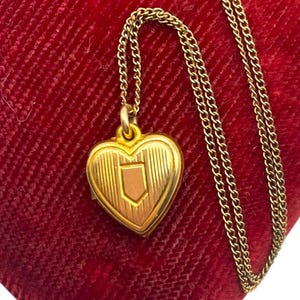 May include: A gold-colored heart-shaped locket pendant with a shield design, suspended from a delicate chain. The locket rests on a red velvet surface, creating a striking contrast. The chain is approximately 18 inches long.