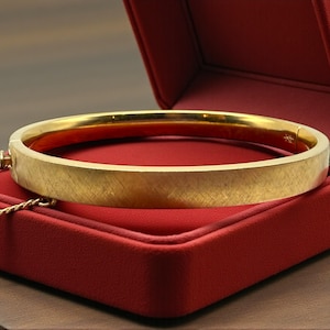 May include: A gold bangle bracelet with a textured surface rests inside a red jewelry box. The bracelet is a solid band of gold, and the box is open, revealing a soft, red interior. A gold chain is draped over the box.
