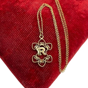 May include: A gold-colored necklace with a pendant featuring the letter "R" in a floral design. The necklace is displayed on a red velvet surface. The chain is delicate and the pendant has intricate details.