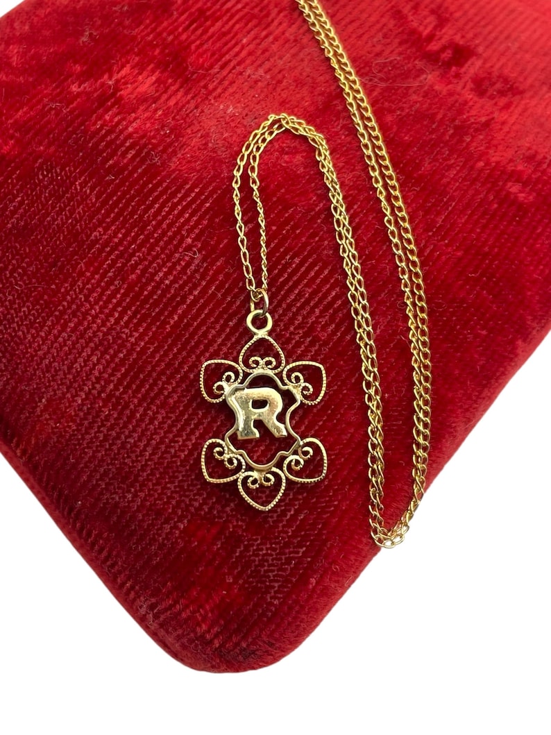 May include: A gold-colored necklace with a pendant featuring the letter "R" in a floral design. The necklace is displayed on a red velvet surface. The chain is delicate and the pendant has intricate details.