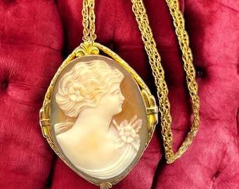 May include: A vintage gold-tone cameo pendant necklace. The oval pendant features a carved profile of a woman with a flower in her hair. The cameo is set in a gold frame and hangs from a long, gold chain. The background is a deep red.