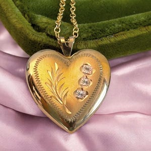 May include: A gold heart-shaped locket pendant on a gold chain. The locket features an engraved floral design and three small square accents. The locket is displayed on a pink satin fabric and a green velvet surface.