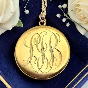 May include: A round gold locket pendant featuring an intricate, hand-engraved monogram with the letters LFB in a flowing script style. The locket is suspended from a matching gold rope chain, showcasing a polished finish and a classic circular shape.