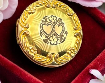 Puede incluir: Un collar con colgante de medallón dorado. El medallón presenta un diseño floral en los bordes y un patrón detallado de corazones y flores en el centro. Se muestra sobre una superficie de terciopelo rojo, con flores rosas de fondo.