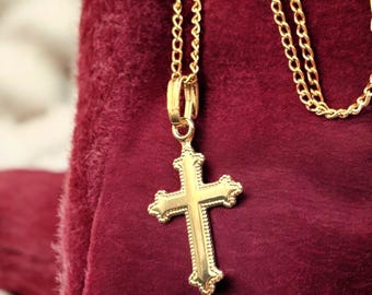 May include: A gold cross pendant necklace with a fine chain. The cross features detailed embellishments and hangs against a deep red backdrop. The necklace is a religious item.