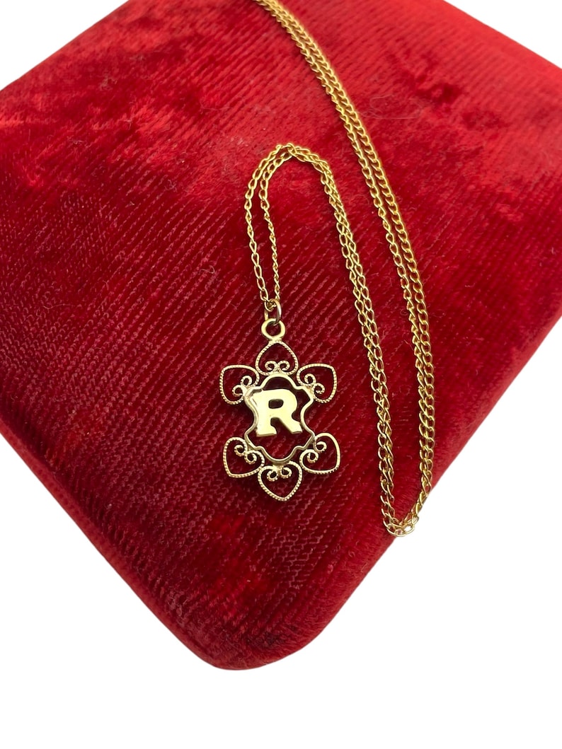 May include: A gold-colored necklace with a pendant featuring the letter "R" in a decorative frame. The pendant is suspended from a delicate chain, resting on a red velvet surface. The necklace is a jewelry item.