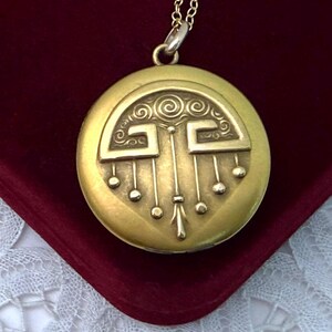 May include: A gold-colored round pendant necklace with an intricate design. The pendant features a stylized pattern with curved lines and small circular accents. The necklace is displayed on a dark red surface.