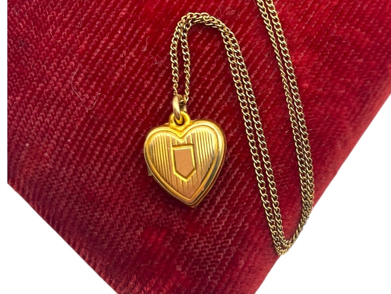 May include: A gold-colored heart-shaped locket pendant with a shield design, suspended from a delicate chain. The locket rests on a red velvet surface, creating a striking contrast. The chain is approximately 18 inches long.