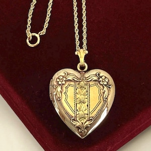 May include: A heart-shaped locket necklace with a gold chain. The locket is silver with gold accents and floral detailing. The center of the locket has a gold shield design. The necklace is displayed on a red velvet surface.