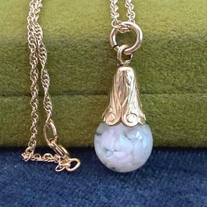 May include: A gold-colored pendant necklace featuring a teardrop-shaped charm filled with white and blue-toned stones. The charm is suspended from a delicate gold chain. The background is a dark blue and green.