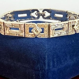 May include: A silver-toned bracelet with a geometric design. The bracelet features rectangular and square links, some of which are embellished with small, clear, and amber-colored rhinestones. The bracelet is displayed on a dark blue velvet surface.