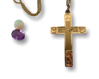 May include: A gold-coloured cross pendant with an engraved design, suspended from a delicate chain. Two gemstones, one purple and one iridescent, are positioned near the chain. The cross has the initials "W.F." engraved on it.