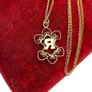 May include: A gold-colored necklace with a pendant featuring the letter "R" in a decorative frame. The pendant is suspended from a delicate chain, resting on a red velvet surface. The necklace is a piece of jewelry.
