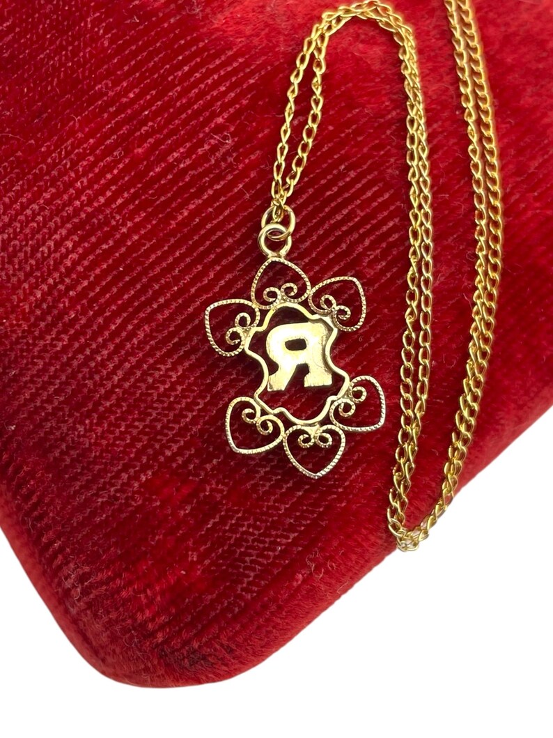 May include: A gold-colored necklace with a pendant featuring the letter "R" in a decorative frame. The pendant is suspended from a delicate chain, resting on a red velvet surface. The necklace is a piece of jewelry.
