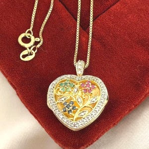 May include: A gold-colored heart-shaped pendant necklace. The pendant features a floral design with green, red, and blue gemstones, surrounded by small clear stones. The necklace is on a red surface.