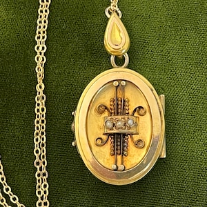 May include: Gold-toned oval locket pendant with a delicate chain. The locket features an ornate design with three small pearls and decorative elements. The pendant hangs on a fine gold chain, set against a green background.