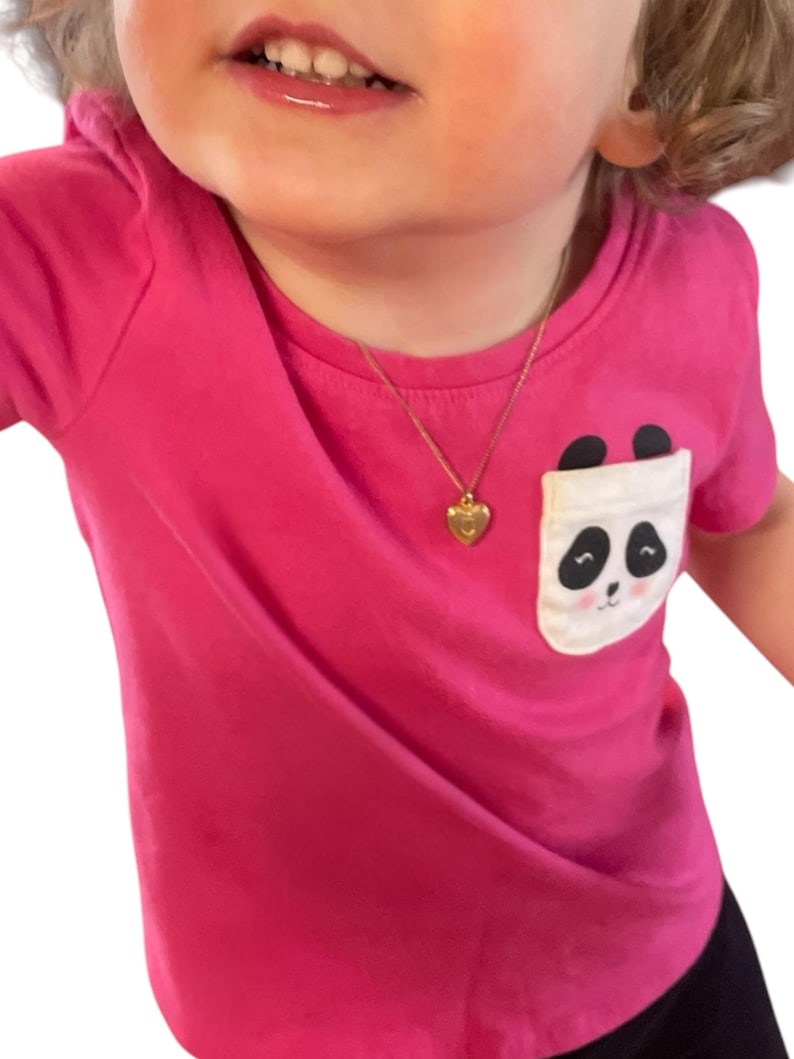 May include: A bright pink t-shirt with a panda bear pocket design. The shirt features a gold heart-shaped pendant necklace. The panda pocket has a white background with a black and white panda face.