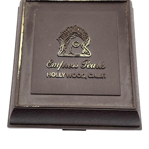 May include: A brown rectangular box with a gold emblem and text that reads "Empress Pearls HOLLYWOOD, CALIF." The box has a raised border and a hinged lid. The emblem is a stylized design.