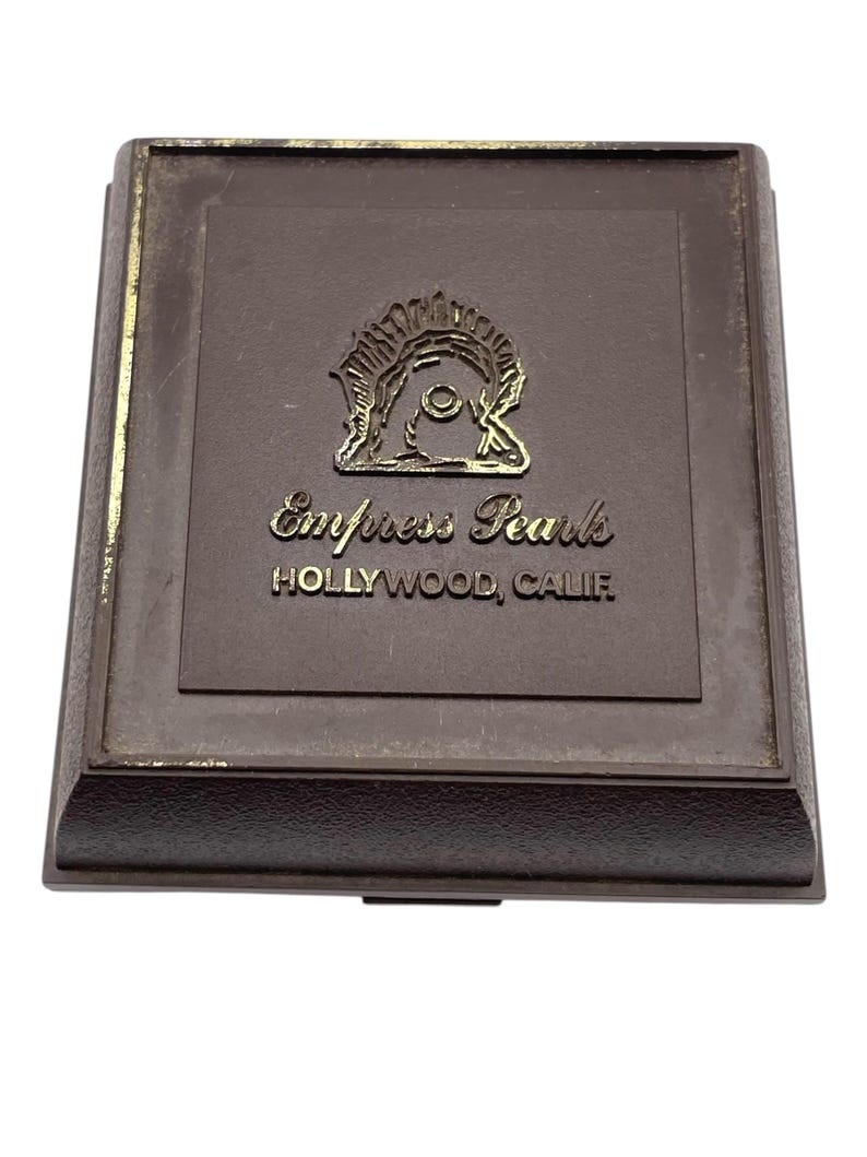 May include: A brown rectangular box with a gold emblem and text that reads "Empress Pearls HOLLYWOOD, CALIF." The box has a raised border and a hinged lid. The emblem is a stylized design.
