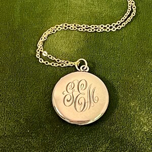 May include: A gold-colored round pendant necklace with an engraved monogram. The pendant hangs from a delicate gold chain. The monogram features the letters "E" and "M" in an elegant script. The necklace rests on a green velvet surface.